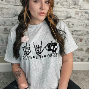 K&ouml;nnte beinhalten: Ein graues T-Shirt mit einem schwarzen Grafikdesign, das Skeletth&auml;nde zeigt, die Friedens-, Rock'n'Roll- und Kaffeenzeichen machen. Der Text "Peace Love Coffee" ist unter dem Grafikdesign gedruckt.