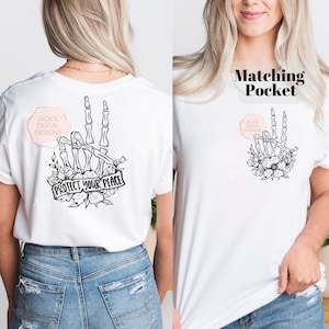 May include: White t-shirt with a black and white skeleton hand peace sign design and floral accents. The text "PROTECT YOUR PEACE" is on a banner. The shirt has a matching pocket with the same design.