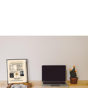 May include: A framed print titled Power of 3-6-9 Cheat Sheet featuring text, a star graphic, and a portrait of Nikola Tesla, displayed next to a silver laptop and a small green cactus in a brown pot on a light wood surface.