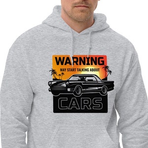 May include: A light gray hoodie with a graphic that reads "WARNING MAY START TALKING ABOUT CARS." The graphic features a black classic car silhouette with palm trees and a sunset background. The hoodie has a drawstring hood and a front pocket.