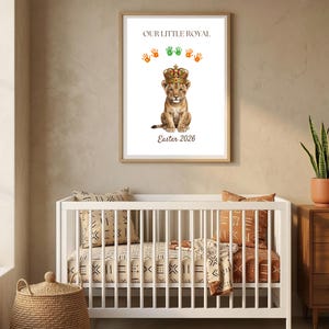 May include: A framed print featuring a watercolor illustration of a lion cub wearing a crown, with the text "OUR LITTLE ROYAL" and "Easter 2026". The print hangs above a white crib with patterned bedding and pillows. A woven basket and a potted plant are also visible.