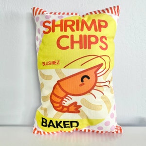 May include: A yellow pillow with a red and white striped border. The pillow has the text "Shrimp Chips Baked" and a cartoon shrimp with a smiling face.