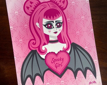 May include: A pink and black illustration of a girl with bat wings and a heart-shaped speech bubble that says "Spooky Girl". The background is a pink and white pattern of skulls and spiderwebs.