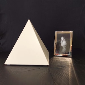 Può includere: Un oggetto decorativo a forma di piramide color crema e una fotografia in bianco e nero incorniciata. La piramide è liscia e geometrica, mentre la fotografia è in una cornice dorata.