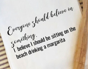 May include: White kitchen towel with black text that reads "Everyone should believe in something. I believe I should be sitting on the beach drinking a margarita."