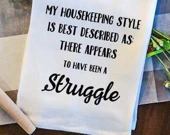 May include: White tea towel with black text that reads "My housekeeping style is best described as: There appears to have been a struggle."