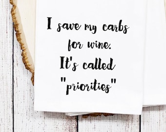 May include: White kitchen towel with the text "I save my carbs for wine. It's called 'priorities'" in black script. The towel is folded and placed on a light wood surface.
