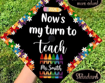 May include: A black graduation cap topper featuring a colorful floral border and the text Now's my turn to teach. It includes a pencil graphic labeled 2026, a row of colorful crayons, and the name Ms. Smith printed in white script lettering.