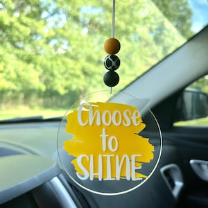 May include: A clear, circular car charm with the words "Choose to SHINE" in white over a yellow paint stroke. The charm hangs from a string with three beads: orange, black and white cow print, and black.