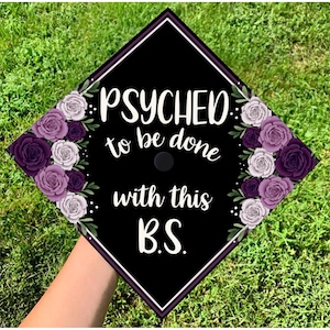 May include: Black graduation cap decorated with purple and white roses. The cap has the phrase "PSYCHED to be done with this B.S." in white script. The cap is held over green grass.
