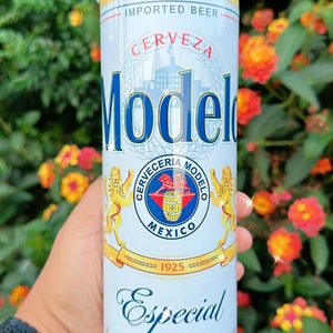 May include: A stainless steel tumbler featuring the Modelo beer logo. The design includes the words "Modelo Especial" and "Cerveza" in blue and gold, with a crest and the words "Imported Beer". The tumbler is held against a backdrop of green foliage and orange flowers.