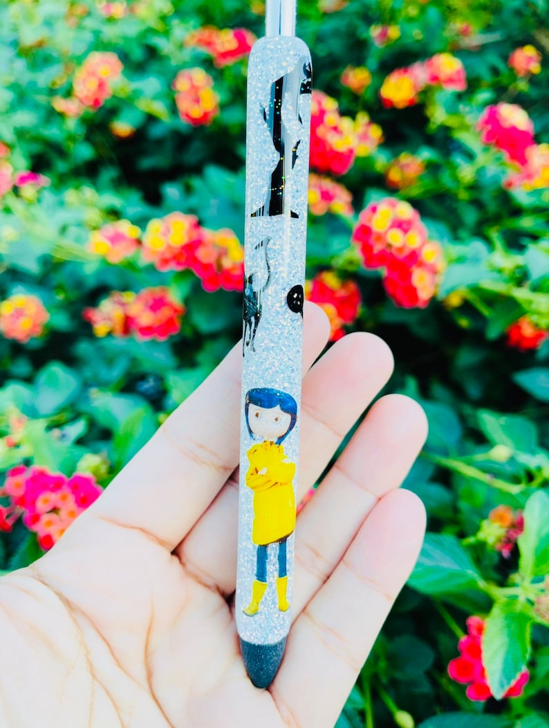 K&ouml;nnte beinhalten: Ein silberner Glitzer-Stift mit einem Schwarzwei&szlig;bild eines M&auml;dchens in einem gelben Regenmantel. Der Stift ist mit Schwarzwei&szlig;bildern von Katzen und einem Mond verziert.