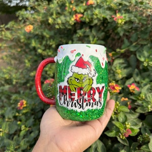 May include: A green glitter Christmas mug featuring the Grinch wearing a Santa hat. The mug has a red glitter handle and the words "Merry Christmas" in red and white. The mug is decorated with white icing and green sprinkles.