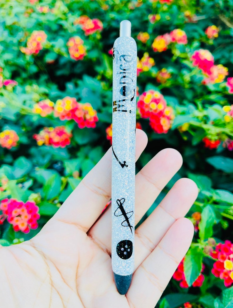 K&ouml;nnte beinhalten: Ein silberner Glitzer-Stift mit schwarzer Tinte und einer schwarzen Kappe. Der Stift hat ein schwarzes Design aus Nadel und Faden mit einem Knopf auf dem Schaft. Der Stift tr&auml;gt die Aufschrift "Monica" auf dem Schaft.
