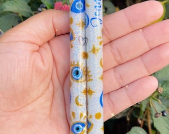 May include: Two white pens with black caps and blue and gold glitter designs. The pens have a pattern of blue evil eyes, gold stars, and moons. One pen has the name "Jennifer" written on it.