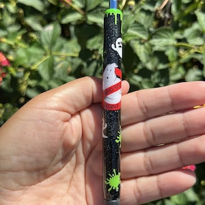 May include: A black glittery retractable pen featuring a white ghost graphic, green slime drip patterns, and a red circular logo with the text WHO YOU GONNA. The pen has a silver click mechanism at the top and a dark gray tip at the bottom.