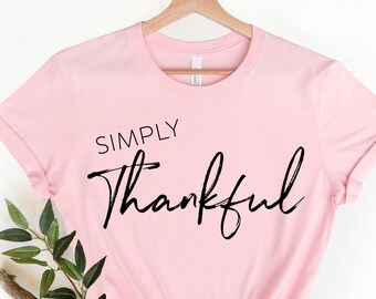 May include: A light pink t-shirt with the words "SIMPLY Thankful" in black script. The shirt is hanging on a wooden hanger. Green leaves and a branch are placed in the lower left corner.
