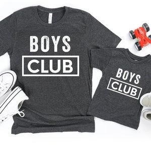 Pode incluir: Duas camisetas cinzas com o texto "Boys Club" em branco. A camiseta maior tem o texto em um tamanho de fonte maior.