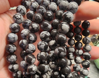 May include: A stack of black and white snowflake obsidian bracelets. Each bracelet is made of round beads with a speckled pattern.