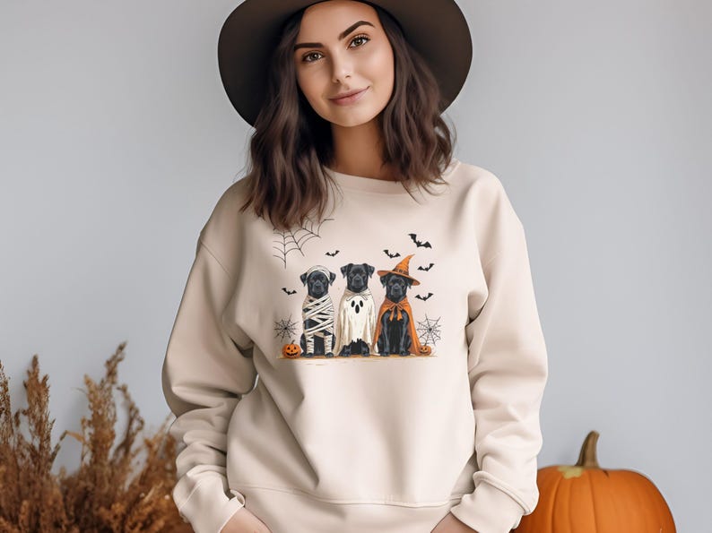 May include: Beige sweatshirt featuring a Halloween-themed graphic. The graphic depicts three black Labrador Retrievers dressed as a mummy, ghost, and witch. Bats, spiderwebs, and pumpkins complete the design. A festive and fun apparel item for the spooky season.