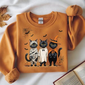 May include: Orange sweatshirt featuring three black cats dressed in Halloween costumes: a mummy, a ghost, and a witch. The design includes bats, spiderwebs, and pumpkins. The Gildan brand tag is visible.