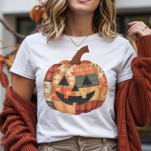 May include: White t-shirt featuring a patchwork pumpkin design. The pumpkin has a smiling face with triangular eyes and a curved mouth. The pumpkin is made of various fabric patterns in orange, red, and green.