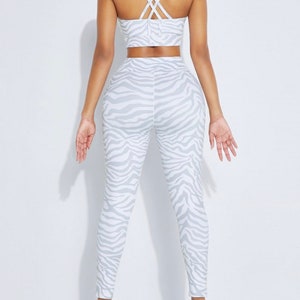 K&ouml;nnte beinhalten: Ein zweiteiliges Sport-Set, bestehend aus einem Sport-BH und einer Leggings. Das Set ist wei&szlig; mit einem grauen Zebra-Streifenmuster. Der Sport-BH hat gekreuzte Tr&auml;ger am R&uuml;cken. Die Leggings sind hoch tailliert.