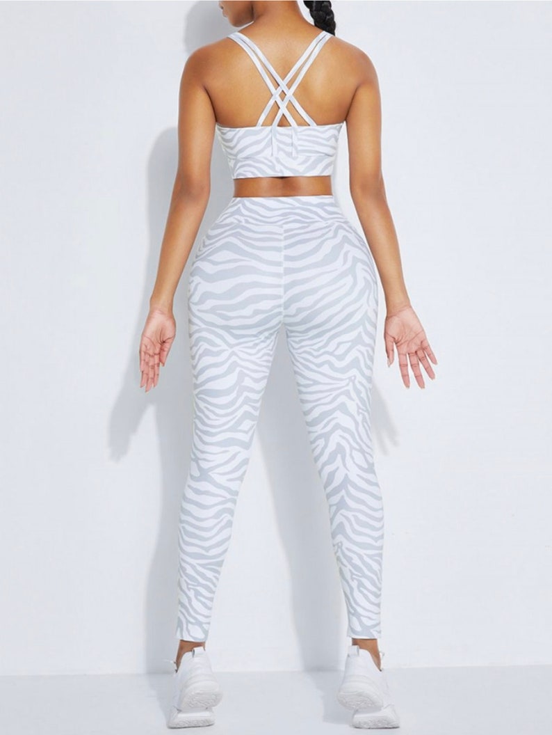 K&ouml;nnte beinhalten: Ein zweiteiliges Sport-Set, bestehend aus einem Sport-BH und einer Leggings. Das Set ist wei&szlig; mit einem grauen Zebra-Streifenmuster. Der Sport-BH hat gekreuzte Tr&auml;ger am R&uuml;cken. Die Leggings sind hoch tailliert.