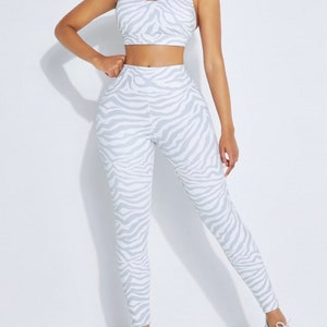 K&ouml;nnte beinhalten: Ein zweiteiliges Sport-Set mit wei&szlig;em und grauem Zebra-Print. Das Set besteht aus einem Sport-BH und einer hoch taillierten Leggings. Das Model tr&auml;gt wei&szlig;e Turnschuhe. Dieses Activewear-Set ist f&uuml;r Workouts und Fitnessaktivit&auml;ten konzipiert.