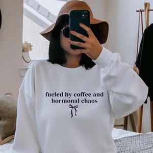 Könnte beinhalten: Weißes Langarm-Sweatshirt mit dem Text "fueled by coffee and hormonal chaos" in schwarzer Schrift. Unter dem Text befindet sich eine schwarze Schleifen-Grafik. Das Sweatshirt hat einen lässigen Stil.