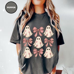 May include: Dark gray t-shirt featuring a design of cartoon ghosts and pink bows. The ghosts have small flower details and the word "PEPPER" is in a circle at the top left. The shirt is a comfortable, casual style.