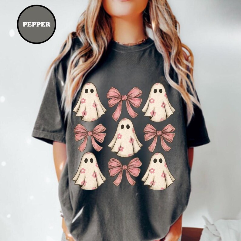 May include: Dark gray t-shirt featuring a design of cartoon ghosts and pink bows. The ghosts have small flower details and the word "PEPPER" is in a circle at the top left. The shirt is a comfortable, casual style.