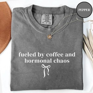 Puede incluir: Una camiseta gris Comfort Colors con la frase blanca "fueled by coffee and hormonal chaos". Un collar plateado con un colgante est&aacute; sobre la camiseta. La camiseta es talla S.