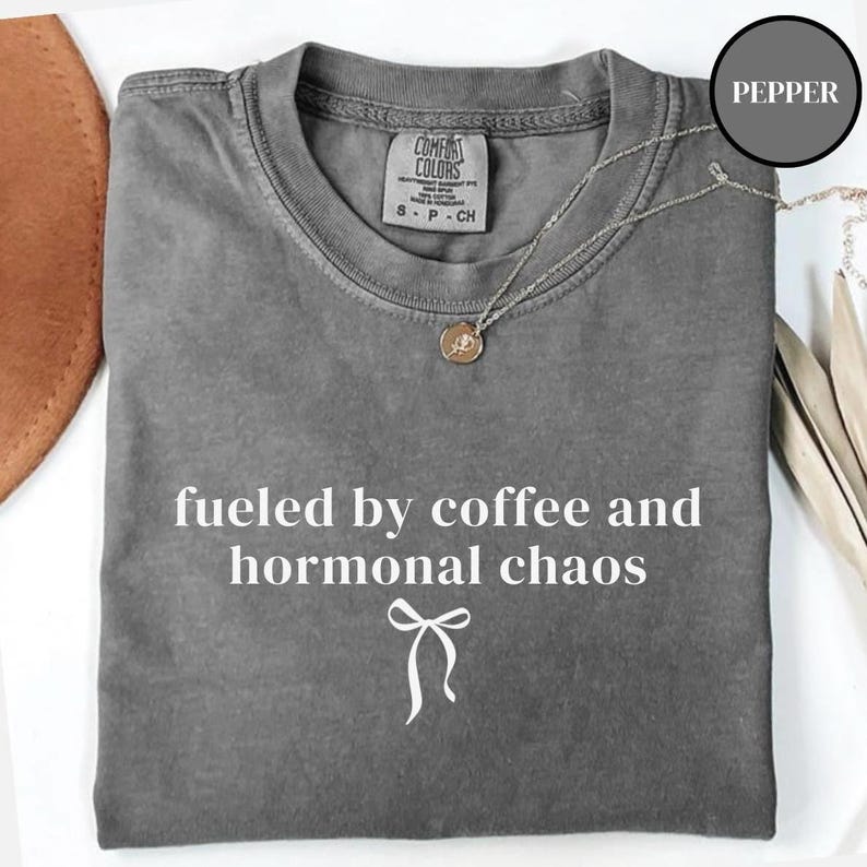 Puede incluir: Una camiseta gris Comfort Colors con la frase blanca "fueled by coffee and hormonal chaos". Un collar plateado con un colgante est&aacute; sobre la camiseta. La camiseta es talla S.