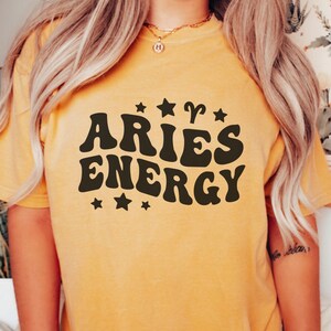 May include: Mustard yellow t-shirt with the words "ARIES ENERGY" in a retro font, surrounded by stars and the Aries zodiac symbol. The shirt is a solid color and has a relaxed fit.