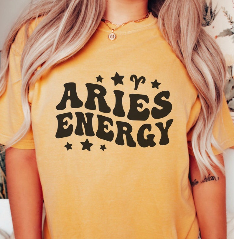 May include: Mustard yellow t-shirt with the words "ARIES ENERGY" in a retro font, surrounded by stars and the Aries zodiac symbol. The shirt is a solid color and has a relaxed fit.