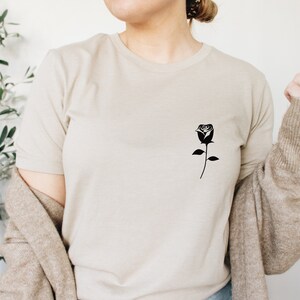 K&ouml;nnte beinhalten: Ein beiges T-Shirt mit einer schwarzen Silhouette einer einzelnen Rose auf der linken Brust.