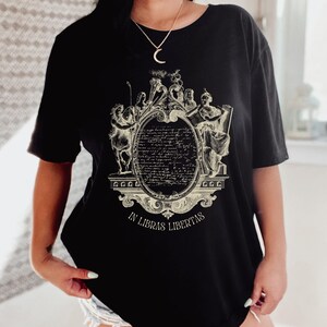 May include: Black t-shirt with a cream-coloured ornate design. The design features figures and text, including an oval filled with script, framed by decorative elements. The text "IN LIBRAS LIBERTAS" is at the bottom of the design.