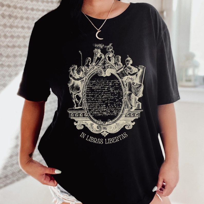 May include: Black t-shirt with a cream-coloured ornate design. The design features figures and text, including an oval filled with script, framed by decorative elements. The text "IN LIBRAS LIBERTAS" is at the bottom of the design.