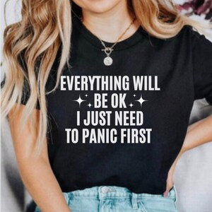 May include: Black t-shirt with the white text "EVERYTHING WILL BE OK I JUST NEED TO PANIC FIRST". The text is centered and includes starburst graphics. The shirt is a crew neck style.
