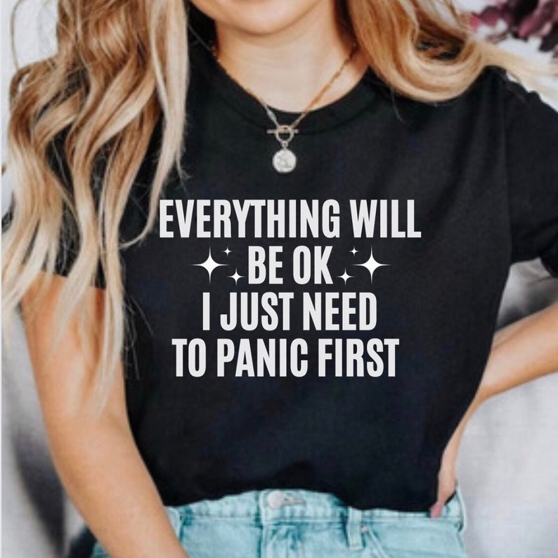 May include: Black t-shirt with the white text "EVERYTHING WILL BE OK I JUST NEED TO PANIC FIRST". The text is centered and includes starburst graphics. The shirt is a crew neck style.