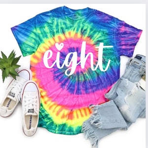Peut inclure: Un t-shirt tie-dye avec un motif en spirale arc-en-ciel. Le t-shirt a le mot "huit" écrit en blanc avec un petit cœur au-dessus du mot.