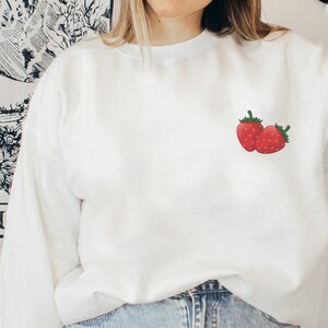 Peut inclure: Sweat-shirt blanc avec un motif de fraise rouge sur la poitrine gauche.