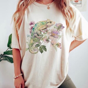 May include: A white t-shirt featuring a green chameleon and pink flowers printed on the front.