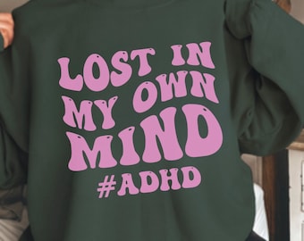 May include: A dark green sweatshirt with the text "Lost in my own mind #ADHD" in pink lettering. The text is in a retro, wavy font.