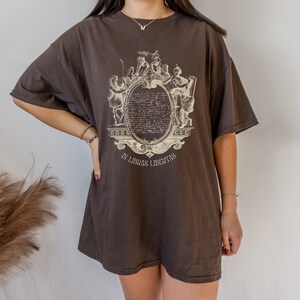 May include: A dark brown oversized t-shirt with a cream-coloured ornate design. The design features figures and text within an oval frame, with the text "IN LIBRAS LIBERTAS". The shirt has short sleeves and a relaxed fit.