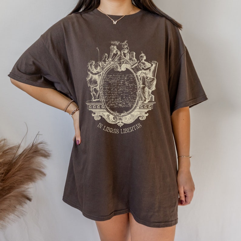 May include: A dark brown oversized t-shirt with a cream-coloured ornate design. The design features figures and text within an oval frame, with the text "IN LIBRAS LIBERTAS". The shirt has short sleeves and a relaxed fit.
