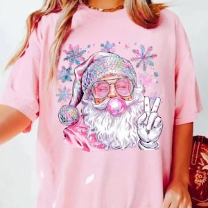 Op de afbeelding: Roze T-shirt met een cartoon afbeelding van de kerstman die een zonnebril draagt en een bel blaast. De kerstman geeft met één hand het vredesteken en houdt in de andere hand een discobal. De achtergrond is een roze aquarelontwerp met sneeuwvlokken.