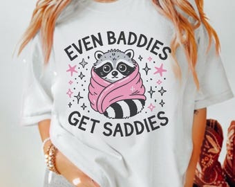 May include: White t-shirt featuring a graphic of a raccoon wrapped in a pink blanket. The text "Even Baddies Get Saddies" is printed on the shirt.