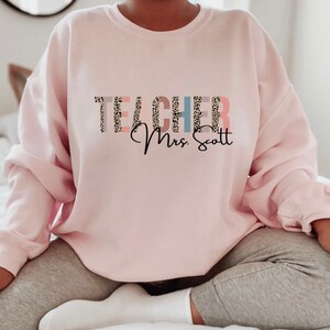 Pode incluir: Camisola cor-de-rosa com a palavra "TEACHER" em estampado de leopardo e o texto "Mrs. Scott" em it&aacute;lico.